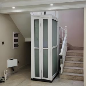 A white residential elevator with glass panels and a white cabin frame located between stairs in a home.