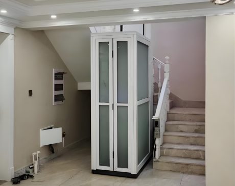 A white residential elevator with glass panels and a white cabin frame located between stairs in a home.