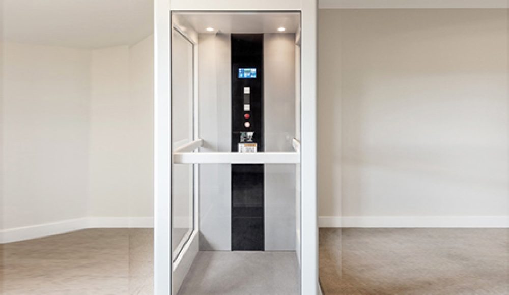 How Much Does A Home Elevator Cost. It's Interior view of a modern home elevator with glass walls and control panel.