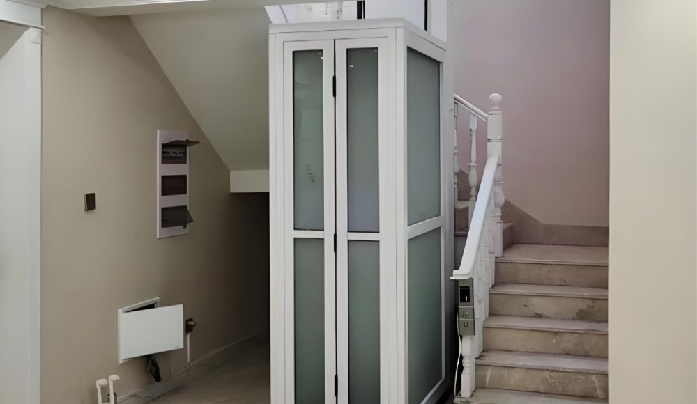 A white residential elevator with glass panels and a white cabin frame located between stairs in a home.